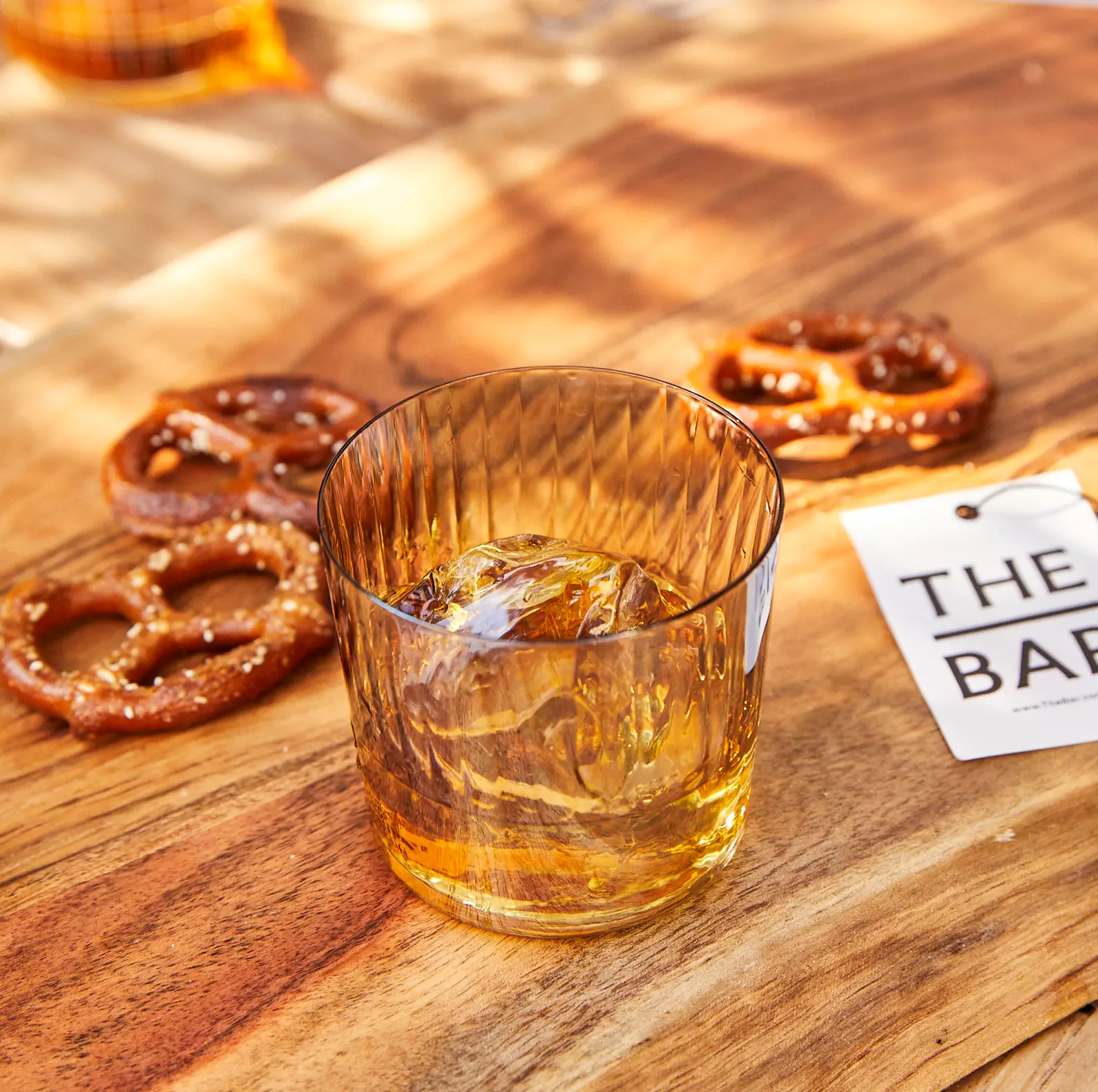 A glass of whiskey with ice sits on a wooden surface next to salted pretzels and a card labeled THE BAR.