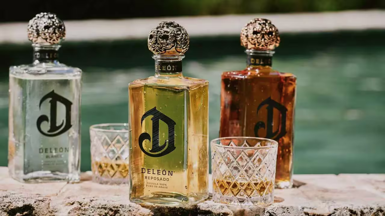 Three bottles of DeLeón tequila and two filled glasses sit on a stone edge beside a pool, with sunlight illuminating the scene.