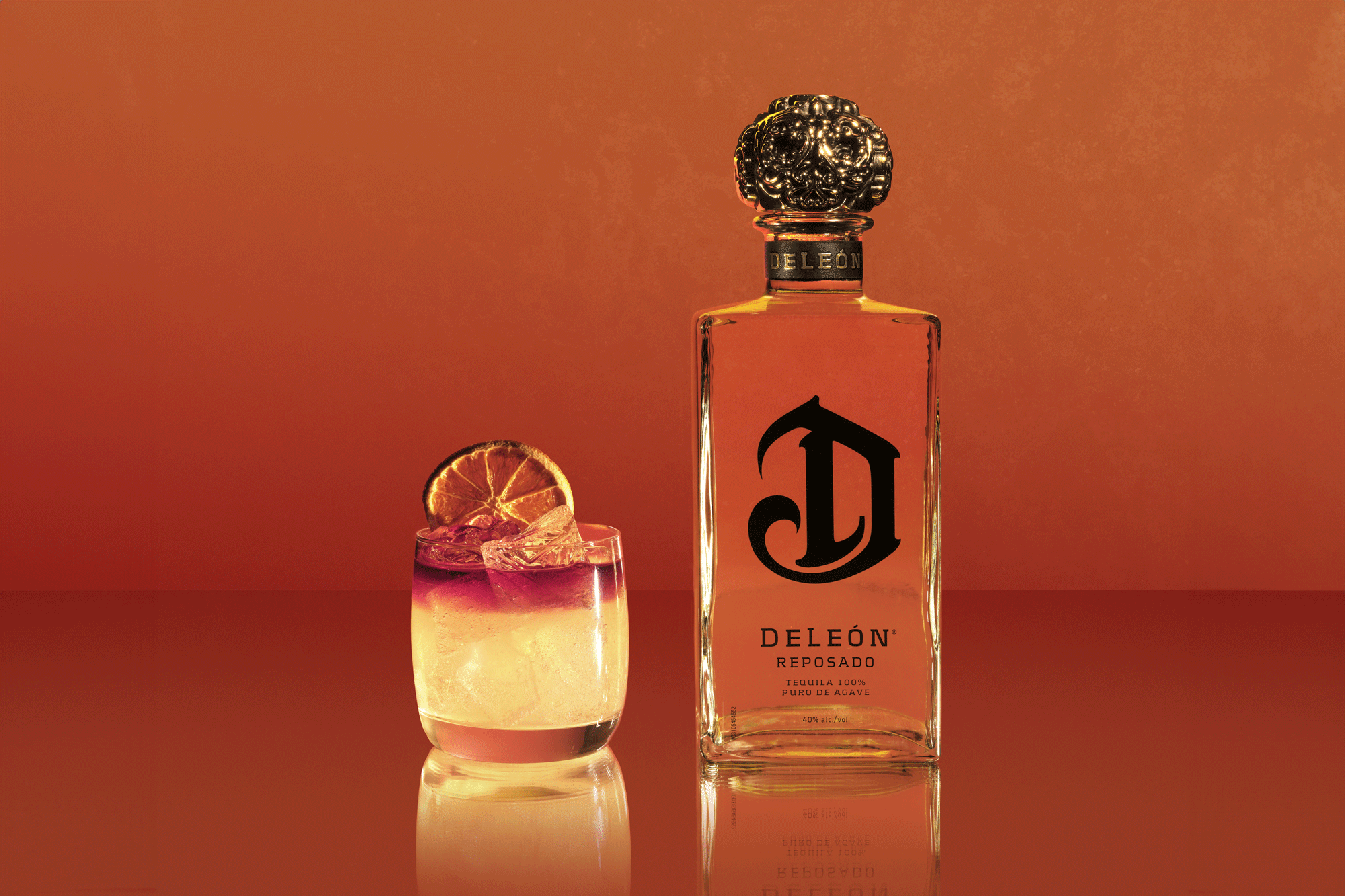A bottle of DeLeón Reposado Tequila stands next to a cocktail with ice and a dried lime slice garnish, set against an orange background.