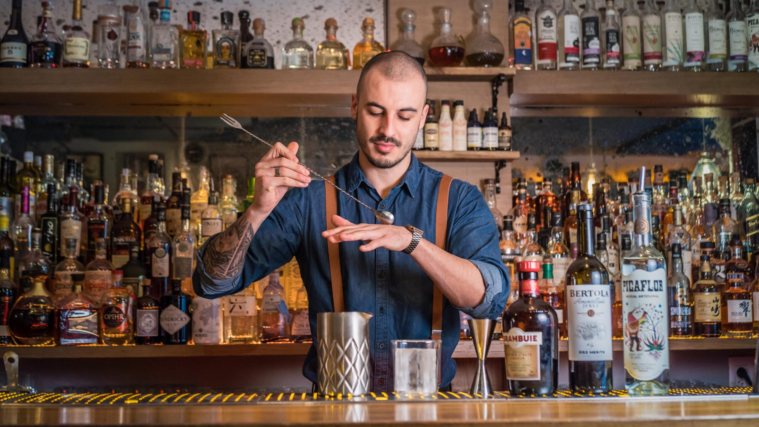 En Bloody Mary Bar, un bartender crea un cóctel con precisión frente a una barra repleta de botellas y detalles cálidos de madera.