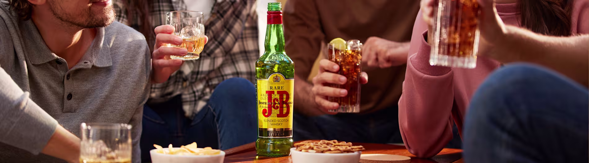 Dos personas se sientan a una mesa puesta sobre hierba con una botella de whisky J&B, un cóctel, platos, cubiertos y pequeños cuencos, y una de ellas sostiene una servilleta roja.