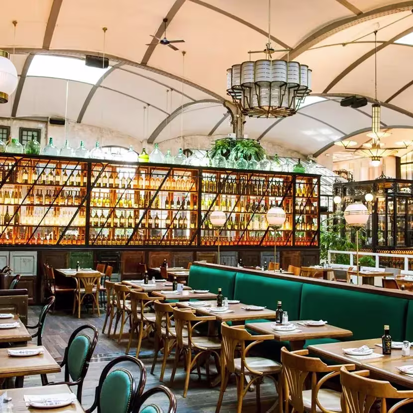 Amplio salón en El Nacional Barcelona, con mesas de madera, sofás verdes, botellas exhibidas y una decoración elegante con plantas colgantes.