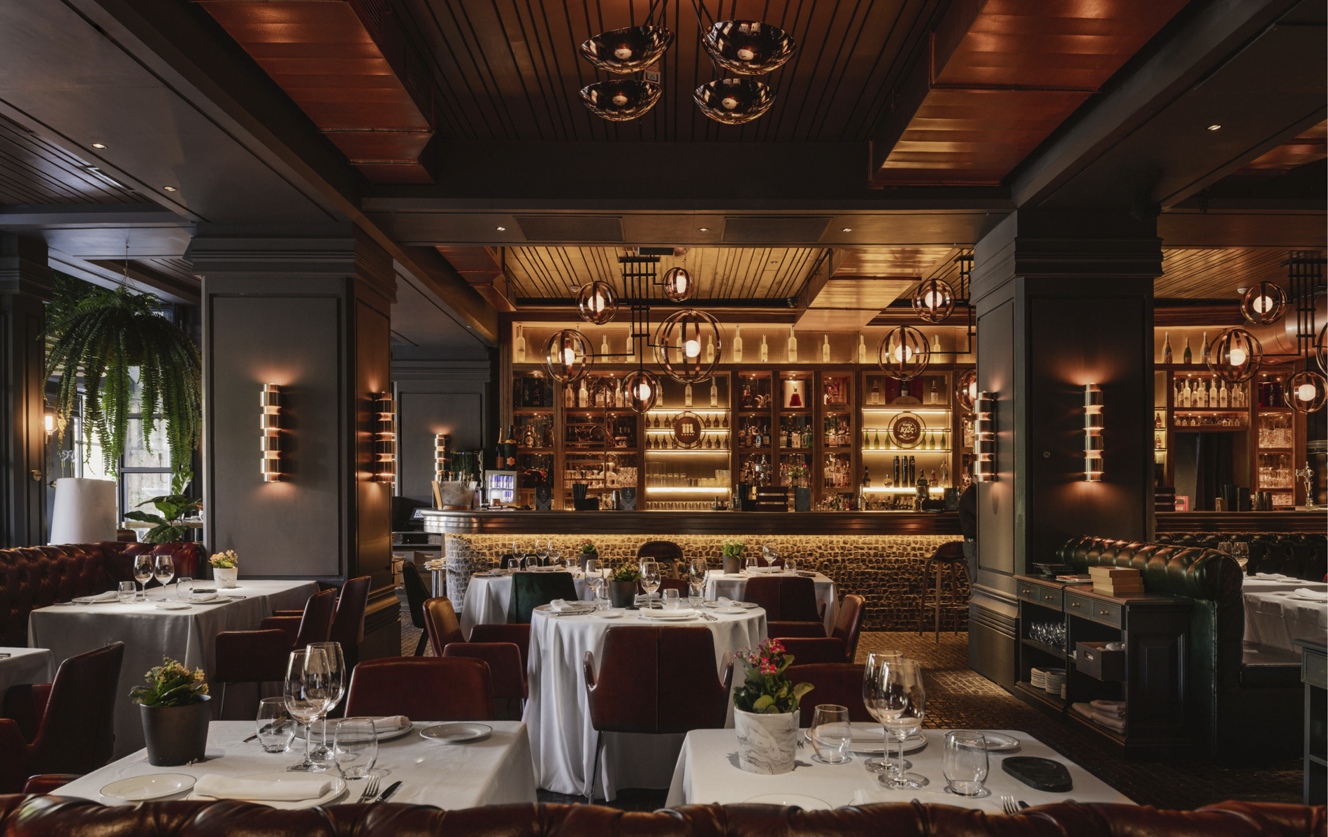 La imagen muestra un elegante comedor del restaurante Tatel. El ambiente es sofisticado, con mesas vestidas con manteles blancos, copas de vino y cubiertos dispuestos ordenadamente. Las paredes y vigas del techo están revestidas de madera oscura.