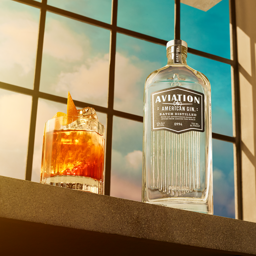 Aviation American Gin bottle next to a cocktail with an orange twist on a windowsill with sunlight streaming through
