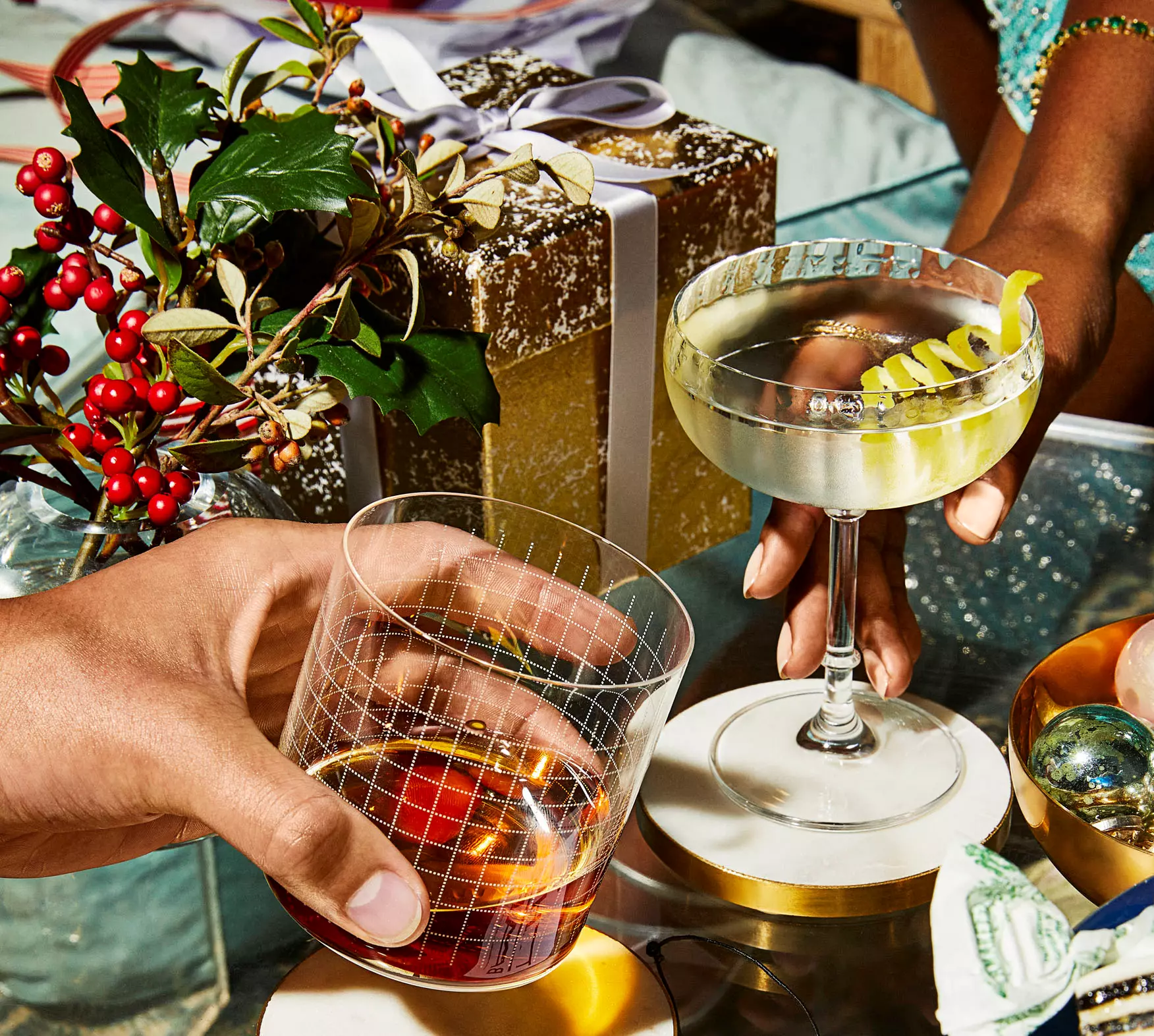Two hands hold cocktails, one in a rocks glass and one in a coupe glass with a lemon twist. A gold-wrapped gift and festive decorations are on the table.