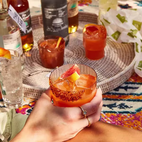 A hand holding a glass of orange cocktail with chili rim and orange garnish; more drinks and bottles on a woven tray in the background.