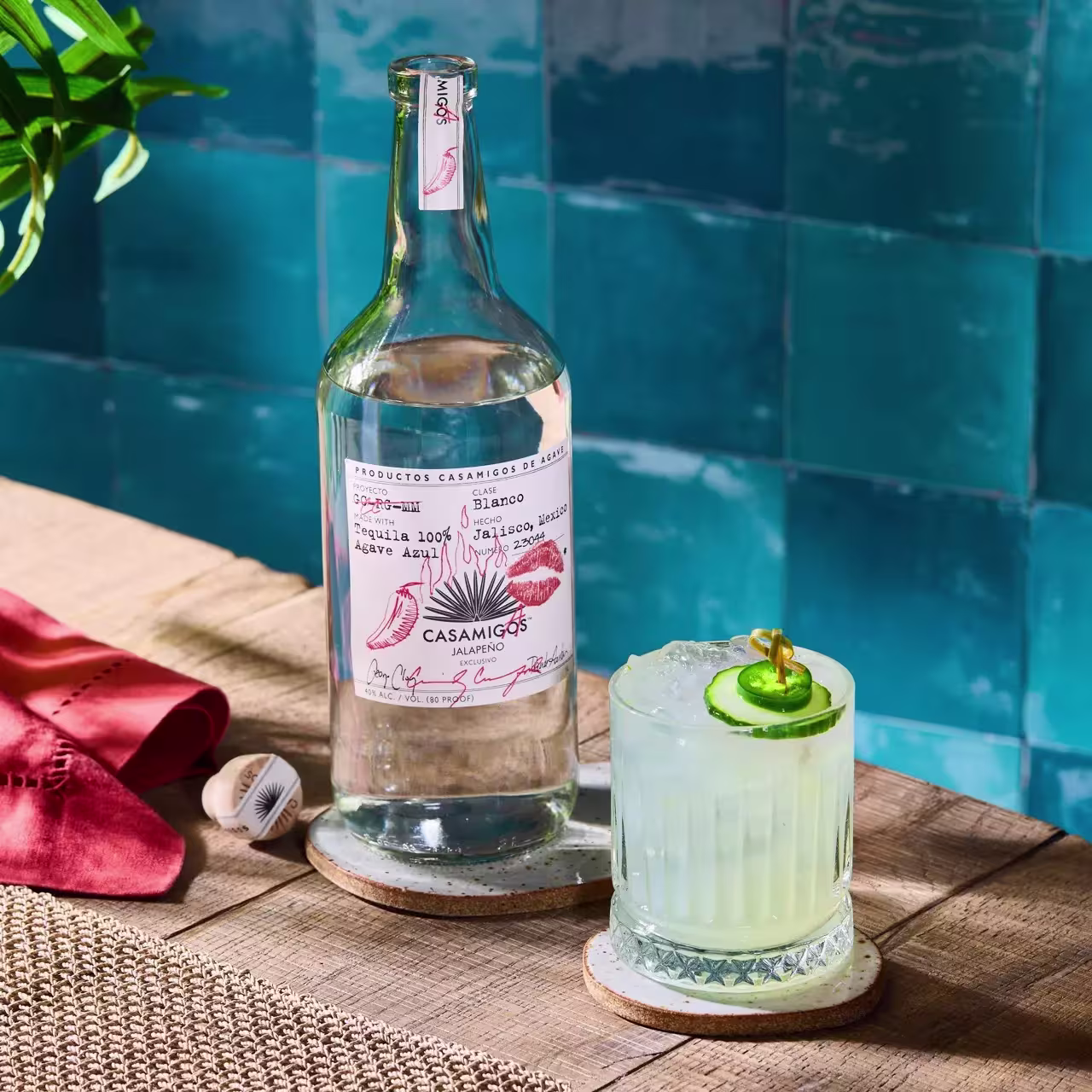 A bottle of Casamigos Blanco tequila and a glass cocktail garnished with cucumber slices sit on a wooden table with a blue tiled wall in the background.