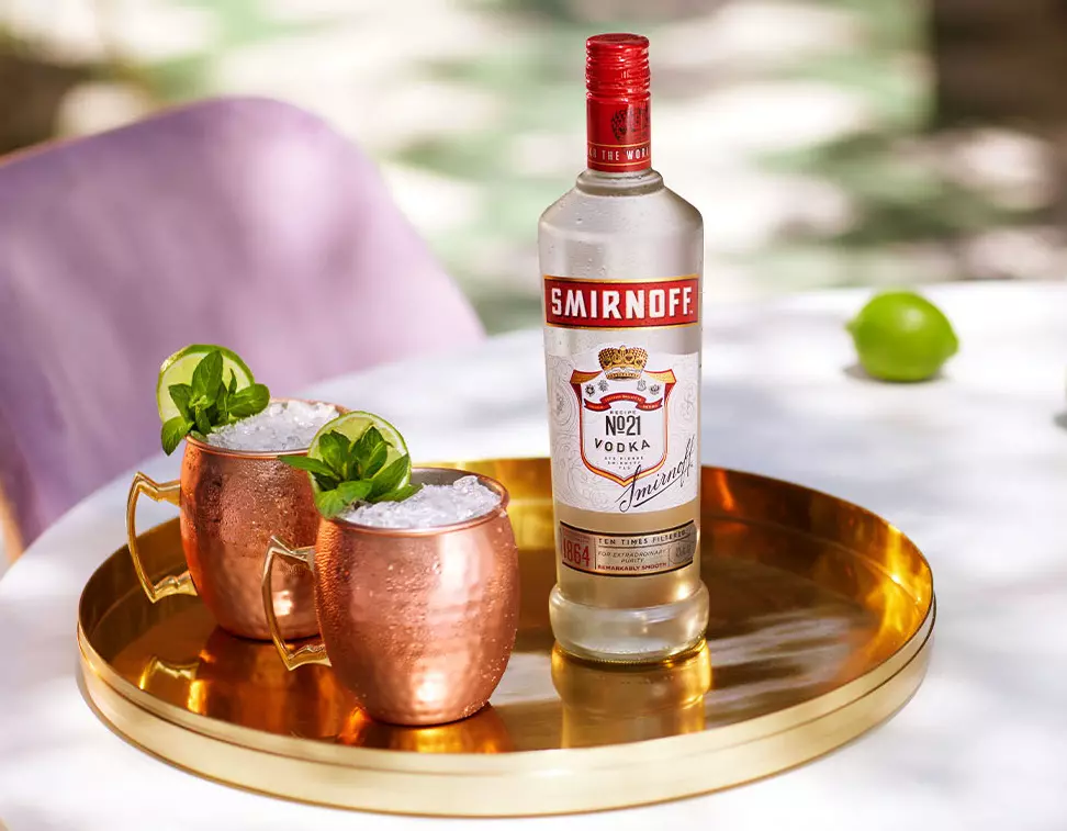A bottle of Smirnoff No. 21 vodka and two copper mugs with ice and garnish sit on a round gold tray, with a lime in the background.
