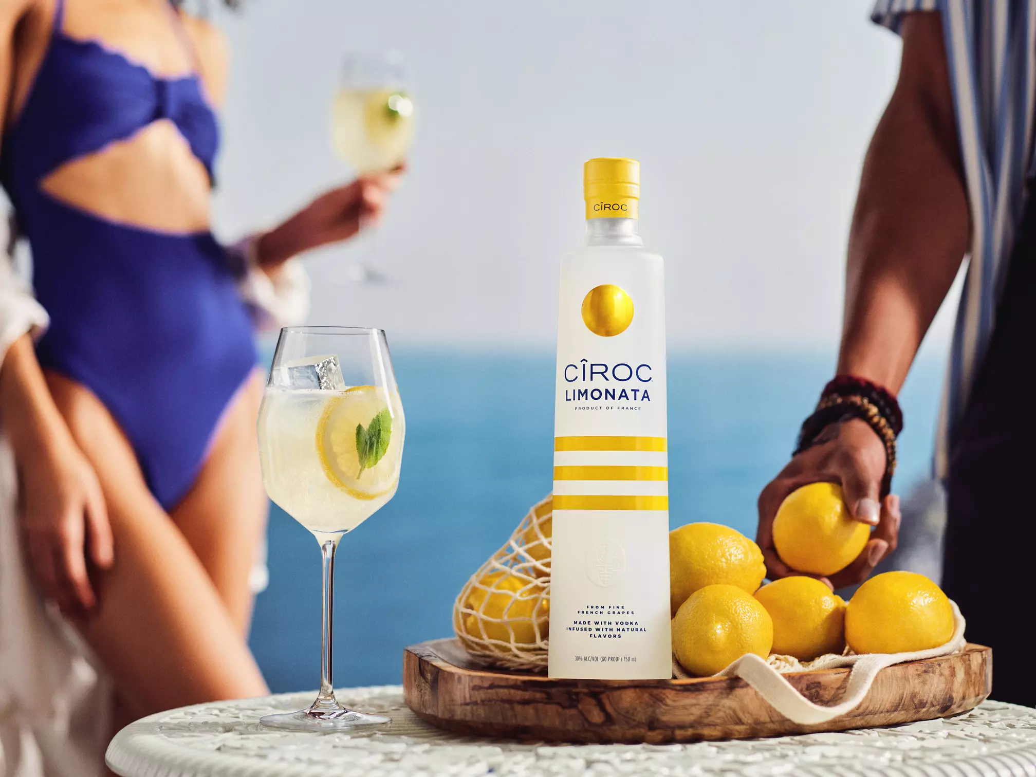 A bottle of Ciroc Limonata stands on a wooden tray with lemons, next to a glass of lemon cocktail; two people are partially visible in the background near water.