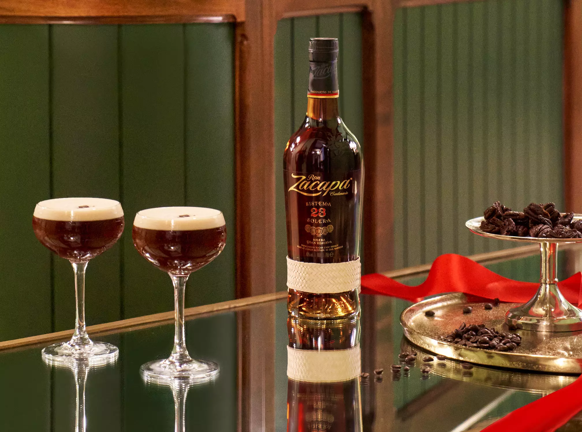 A bottle of Ron Zacapa rum is placed on a reflective table with two espresso martini cocktails, coffee beans, chocolate pieces, and a red ribbon nearby.