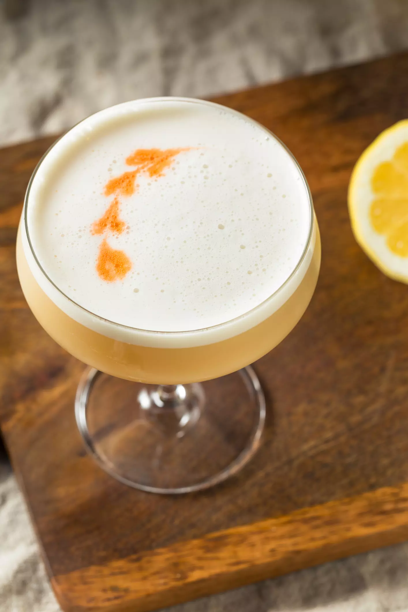 A cocktail in a coupe glass with a foamy top and bitters garnish, placed on a wooden surface next to a lemon slice.