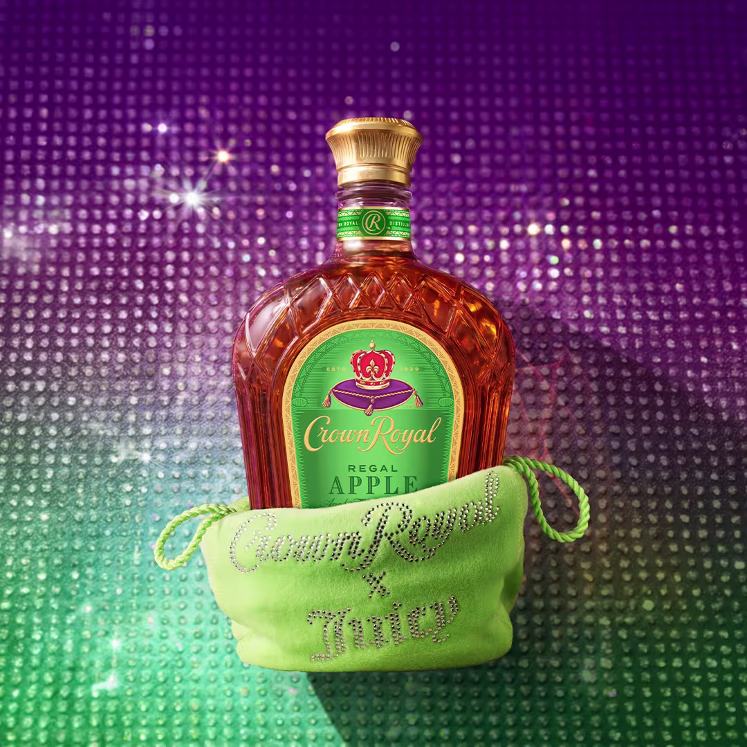 Crown Royal Regal Apple whisky bottle displayed behind a bright green Juicy Couture pouch embellished with rhinestones, set against a sparkling purple and green backdrop.