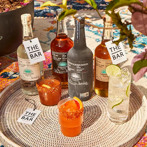 Four bottles of Casamigos tequila and three cocktails with garnishes are arranged on a woven tray atop a colorful rug, with tags reading THE BAR attached to the bottles and tray.