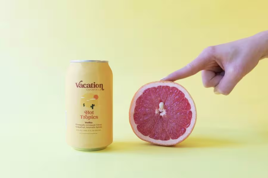 A can labeled Vacation Cocktail Hot Tropics stands next to a halved grapefruit on a yellow background, with a hand pointing at the grapefruit.
