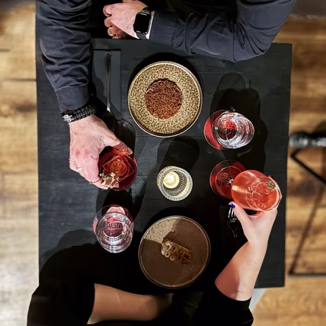 Vista cenital de una mesa con platos de autor y cócteles rosados en copas elegantes; dos personas brindan en un ambiente íntimo y moderno.