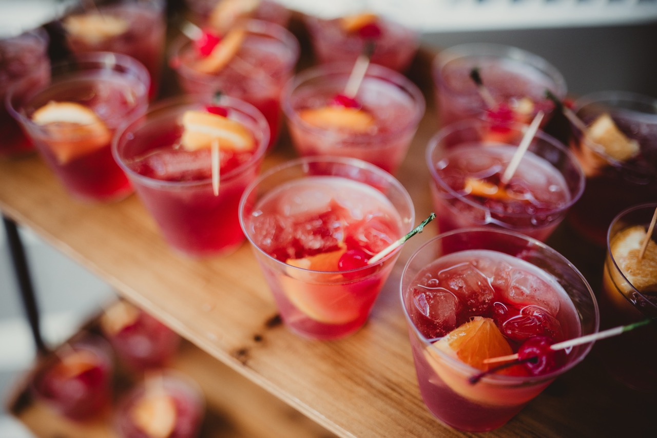 Multiple clear plastic cups filled with red iced drinks, garnished with orange slices and cherries, are arranged on a wooden surface.