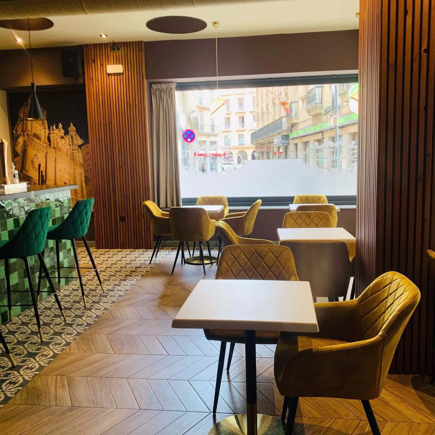 La imagen muestra el interior de The New Salón del Café, con una elegante decoración que combina sillas acolchadas en tonos mostaza, suelos de madera en espiga y una barra de azulejos verdes.