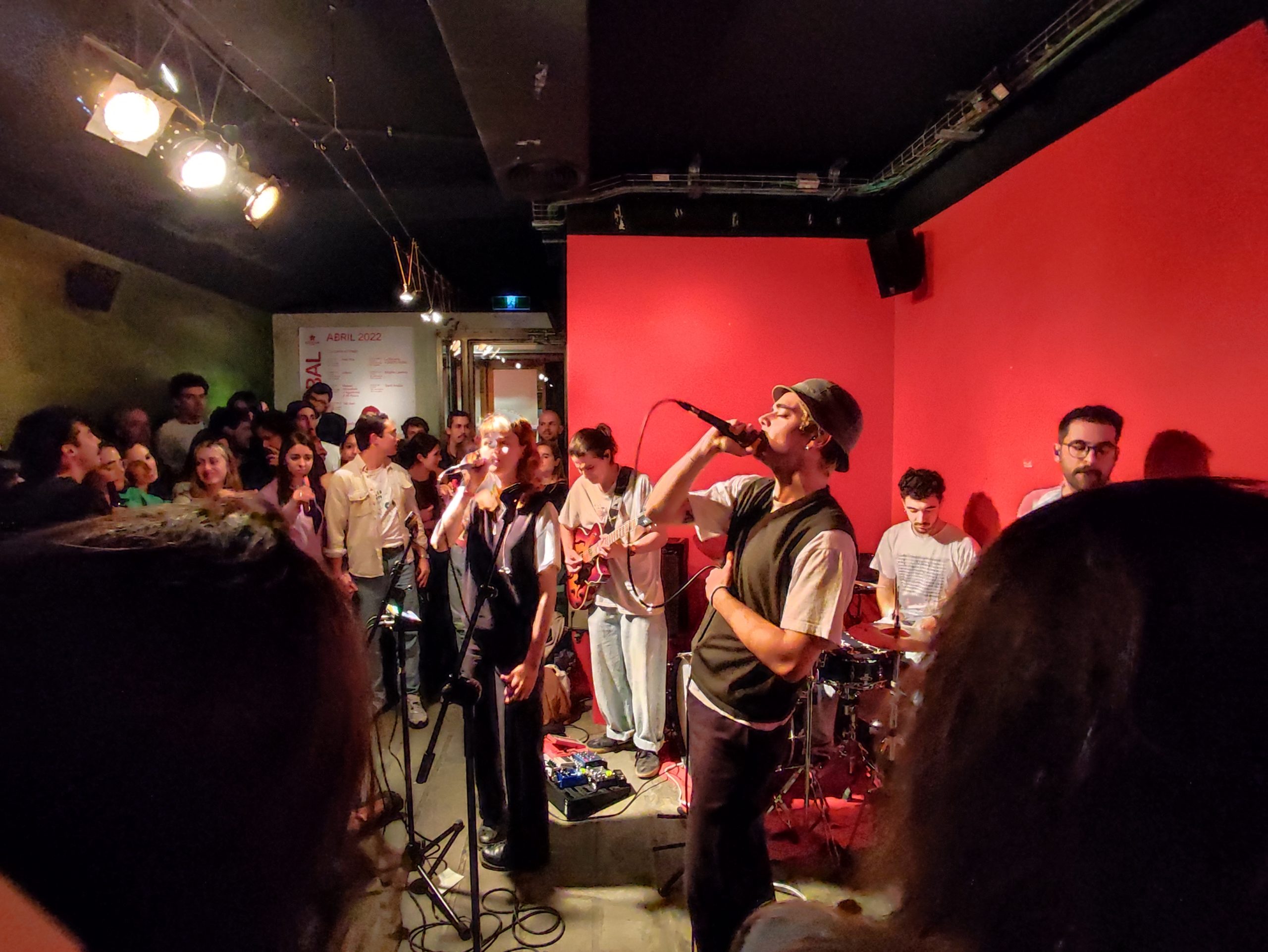 La imagen muestra una presentación en vivo en Heliogàbal, con una banda actuando en un ambiente íntimo. El vocalista y los músicos tocan frente a una audiencia cercana, creando una atmósfera acogedora y vibrante en este espacio de paredes rojas.