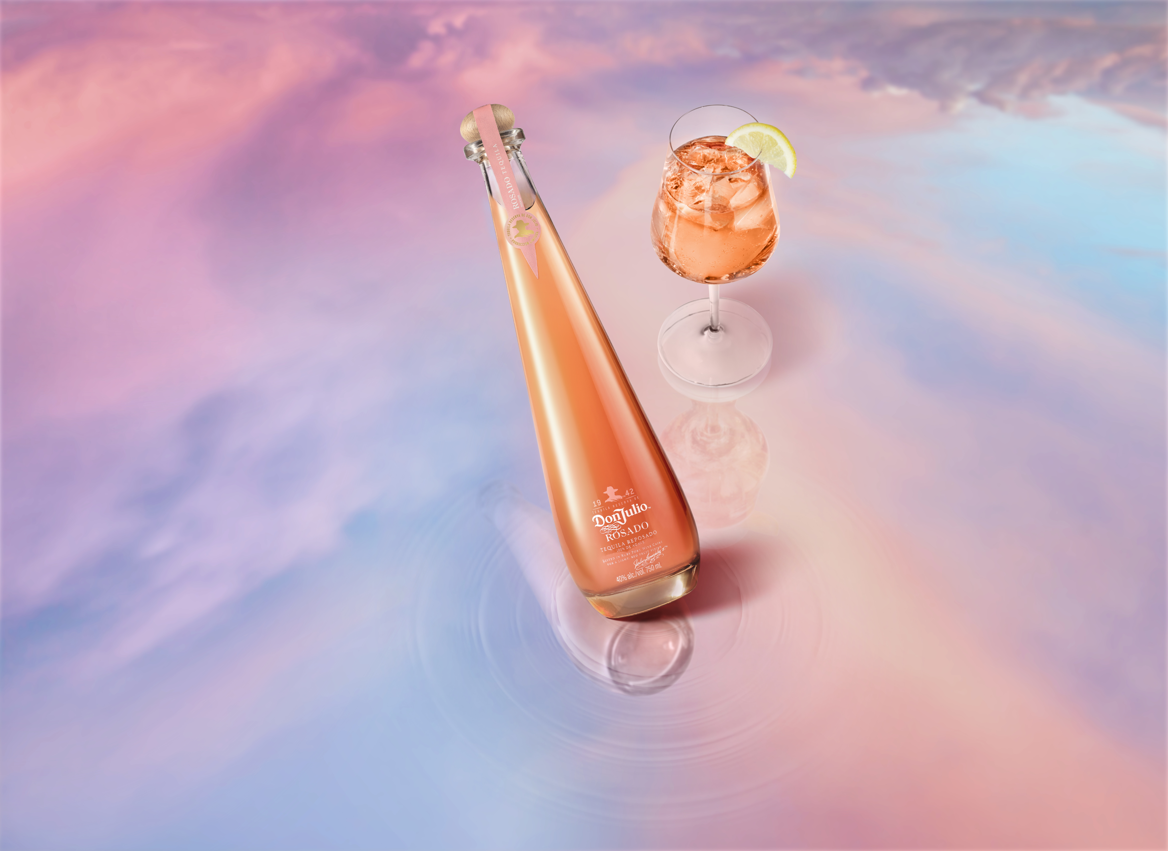 A tall, elegant bottle of Don Julio Rosado tequila stands next to a pink cocktail in a stemmed glass with ice and a lime wedge, set against a pastel-colored reflective surface.