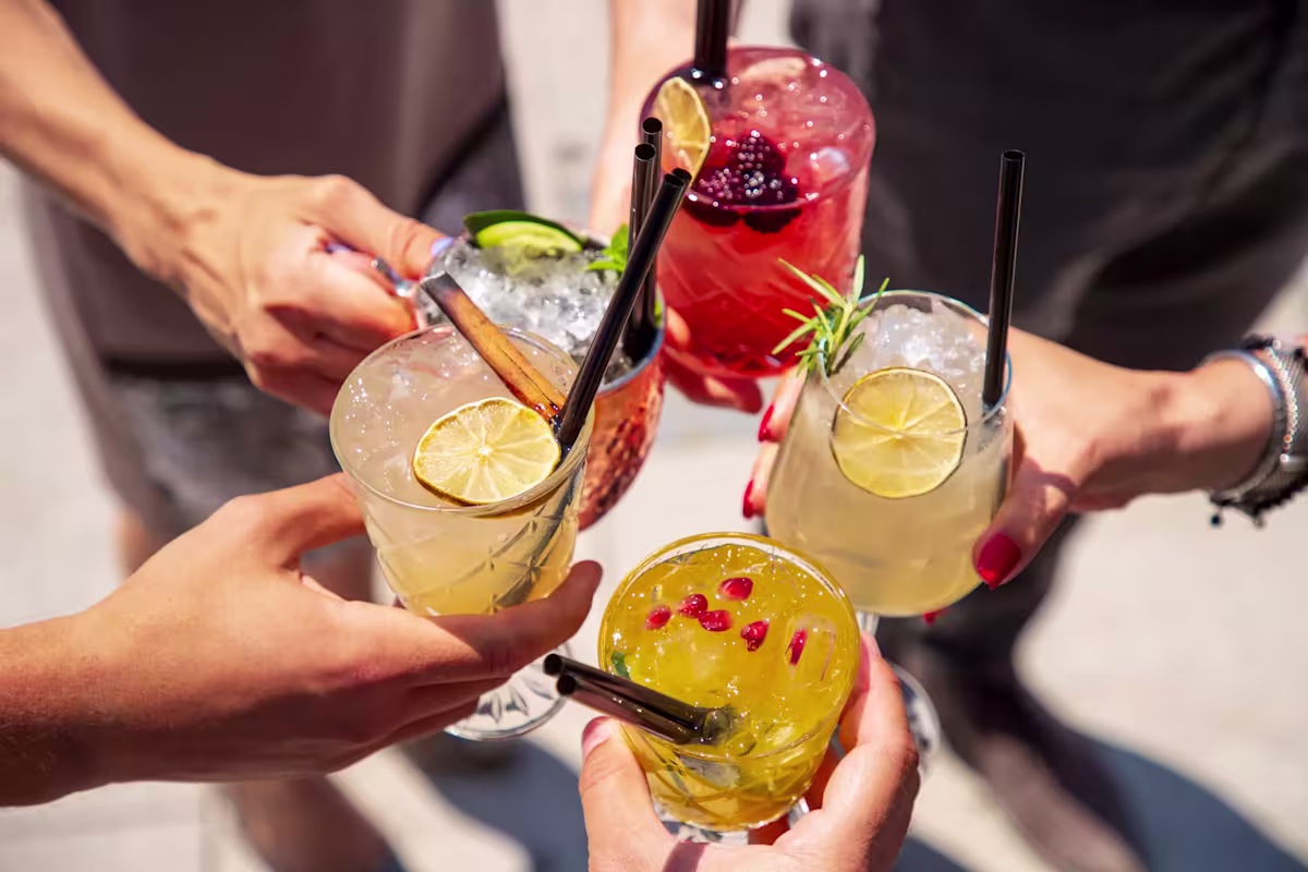 Five people holding various cocktails with ice, fruit garnishes, and straws, toasting together outdoors in daylight.