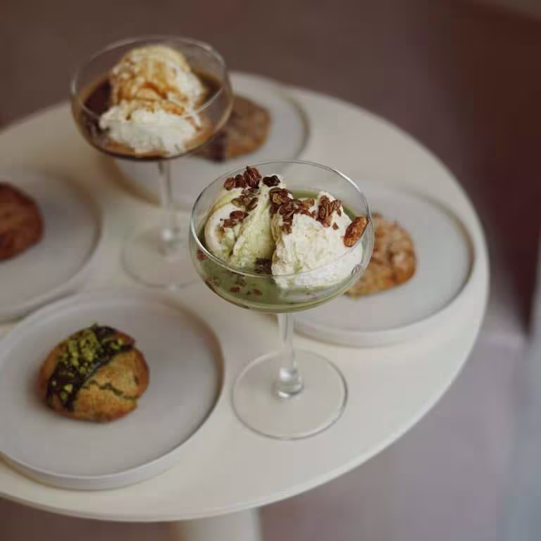 Postres servidos en copas con helado, sirope y frutos secos, acompañados de galletas y bollería sobre platos individuales en una mesa redonda blanca.