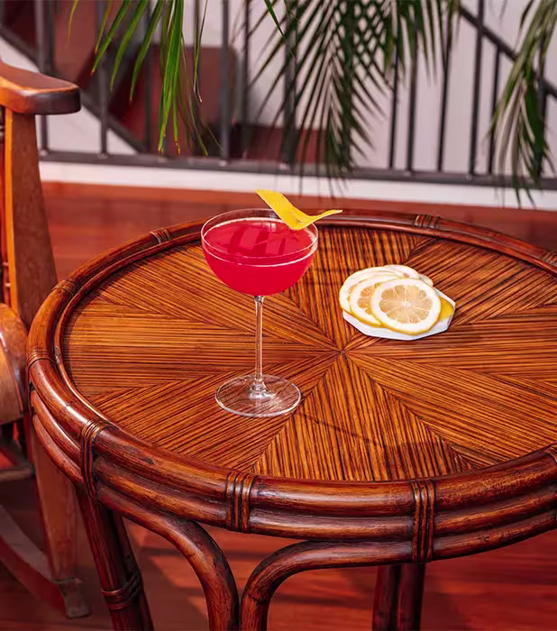 A red cocktail with a lemon twist sits on a round wooden table next to a small stack of lemon slices, with a chair and plants in the background.