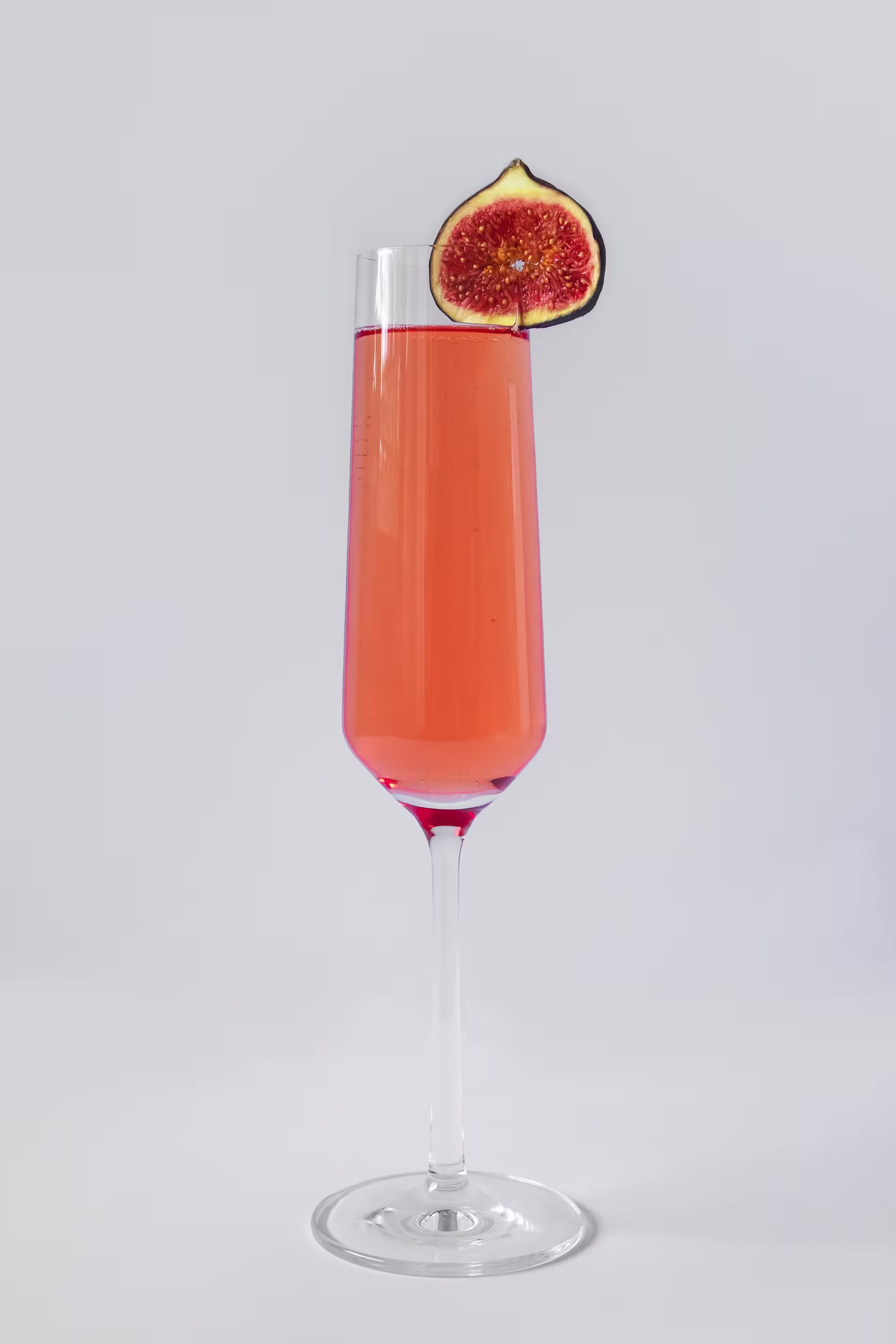 A tall champagne flute with a pink drink, garnished with a slice of fig on the rim, set against a plain light background.