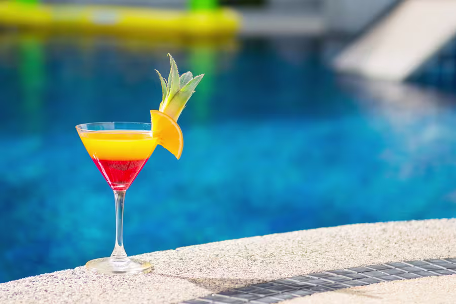 A layered red and yellow cocktail garnished with pineapple and orange slices sits on the edge of a pool with blue water in the background.