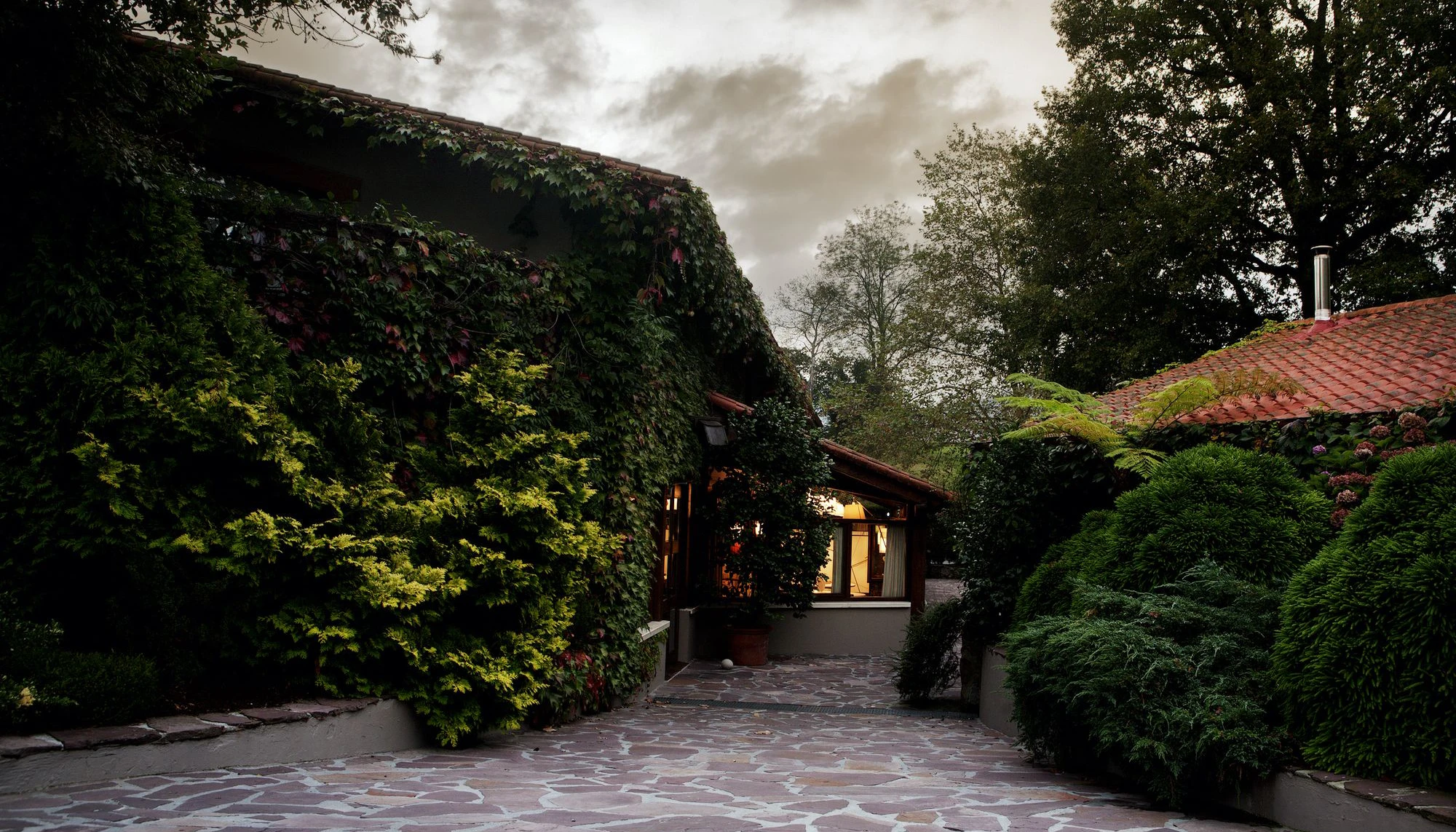 Exterior de Mugaritz, rodeado de abundante vegetación, fachadas cubiertas de enredaderas y un ambiente tranquilo bajo un cielo nublado.