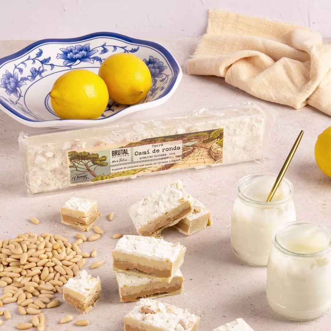 Trozos de turrón "Camí de Ronda" con piñones, limón y azúcar glas, presentados junto a piñones crudos, limones frescos, yogur y una fuente decorativa sobre fondo claro.