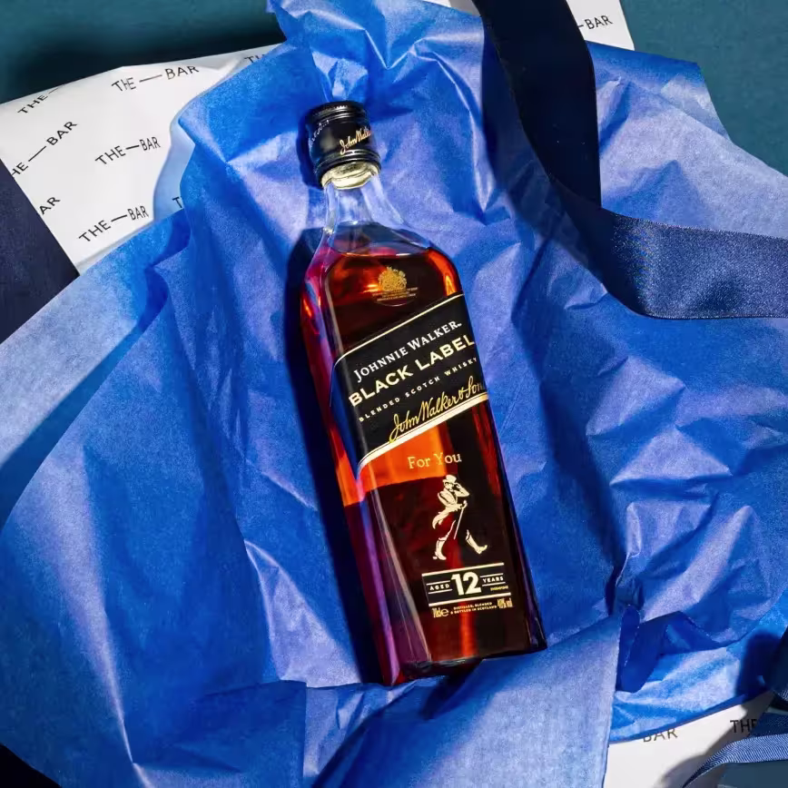 Una botella de whisky escocés Johnnie Walker Black Label 12 años descansa sobre papel de seda azul dentro de una bolsa de regalo.