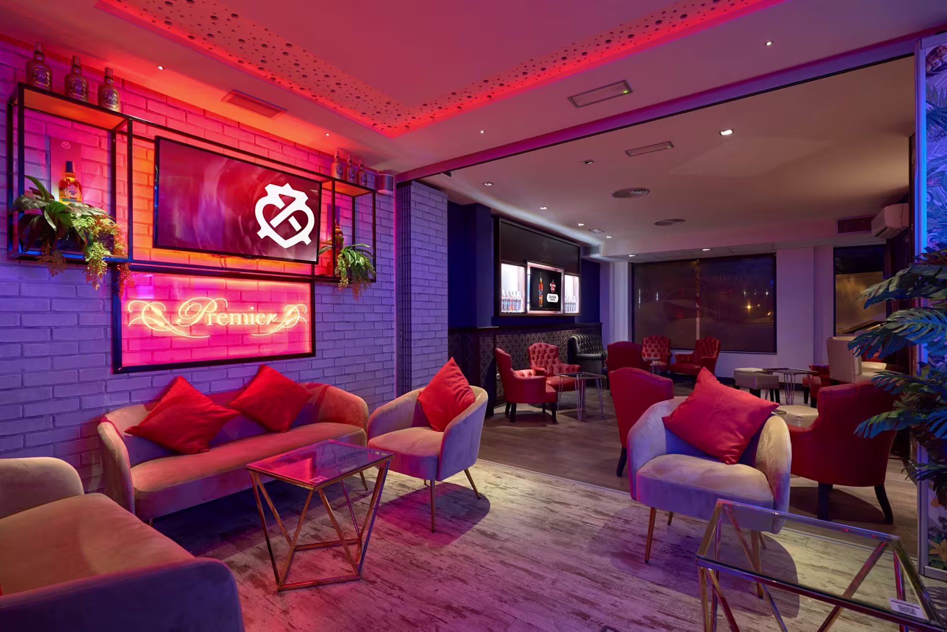 Zona lounge de Premier Cocktail con sofás beige y rojos, luces de neón y decoración moderna. Ambiente elegante y acogedor.