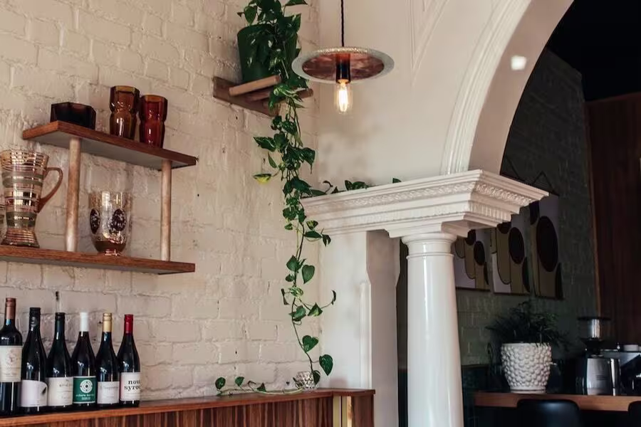 A white brick wall with shelves holding glassware, a row of wine bottles, a hanging plant, and an arched architectural column under a hanging light bulb.