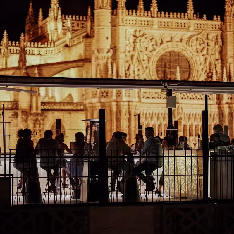 La Terraza del EME al anochecer, con siluetas de comensales y la Catedral de Sevilla iluminada como telón de fondo.