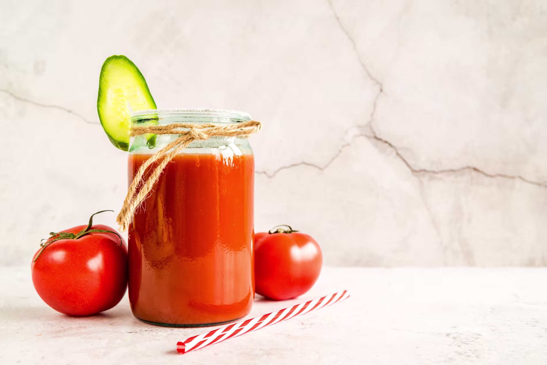 A glass jar filled with tomato juice, garnished with a cucumber slice and twine, sits next to two tomatoes and a red-striped straw on a light surface.