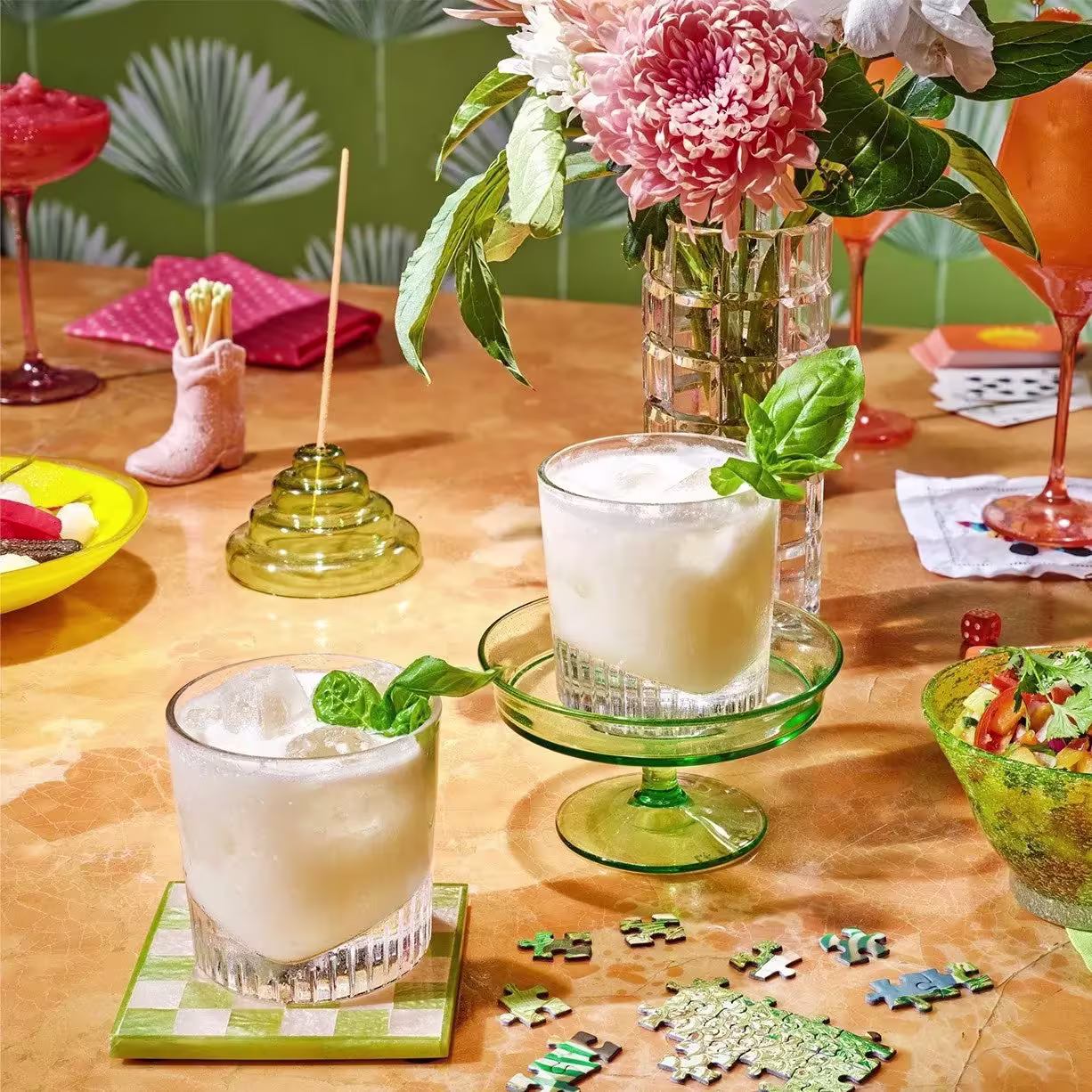 Two frothy cocktails with basil garnish sit on a table with a puzzle, flowers, glassware, and snacks in a colorful, decorative setting.