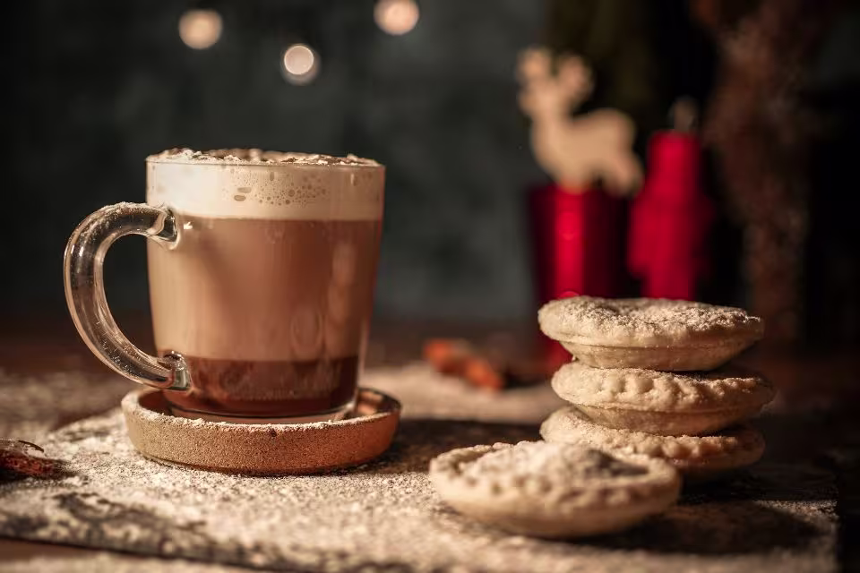 A glass mug of frothy coffee sits on a saucer next to a stack of four powdered pastries on a textured surface, with festive decorations blurred in the background.