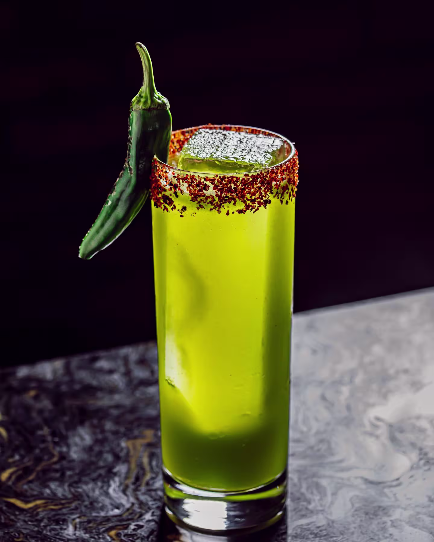 Cóctel verde en vaso alto con borde de chile en polvo, hielo grande y decoración de chile fresco, sobre mesa oscura de mármol.