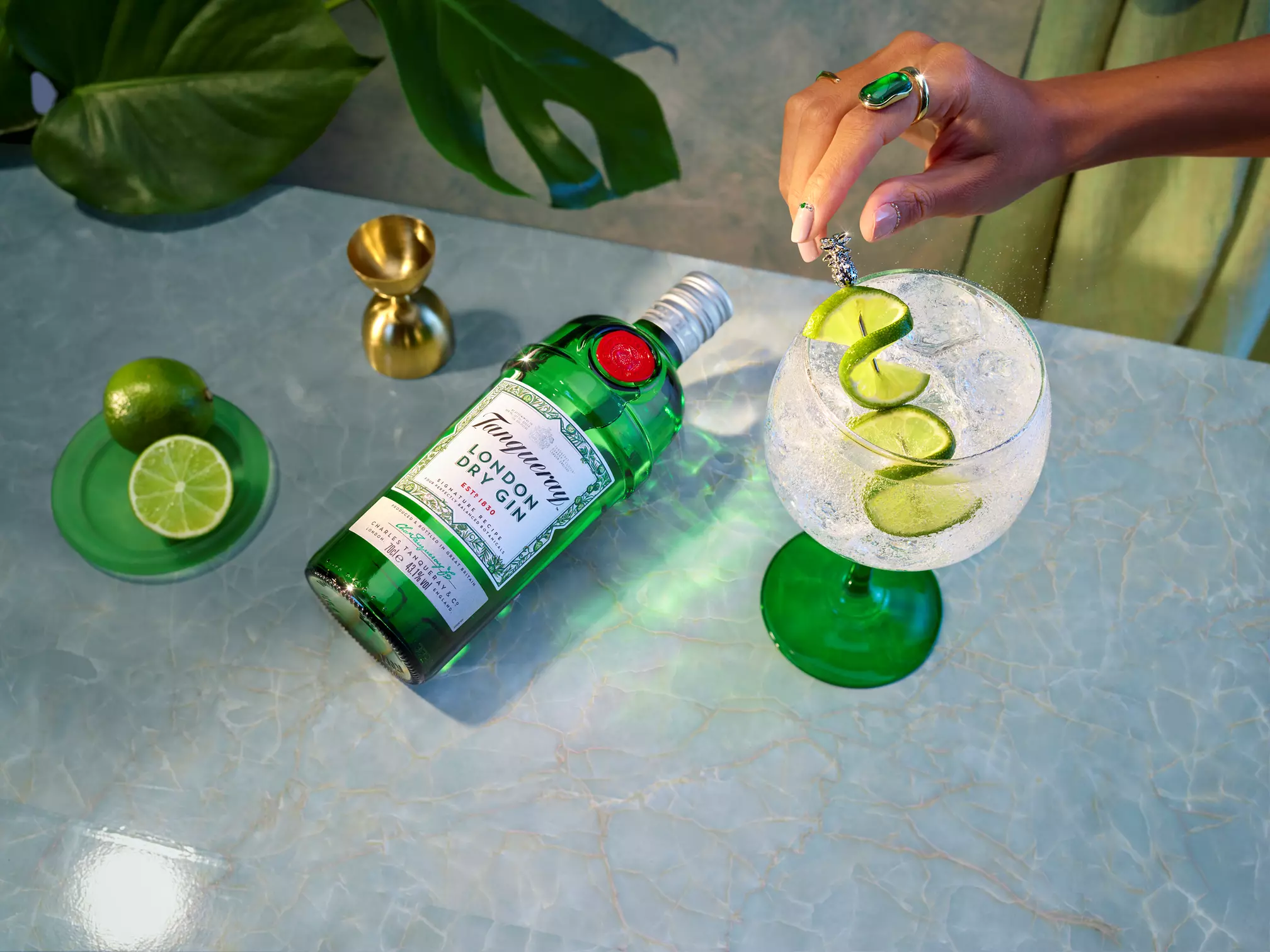 A hand garnishes a gin and tonic with a spiral lime peel beside a bottle of Tanqueray London Dry Gin, a sliced lime, and a jigger on a light blue marble surface.