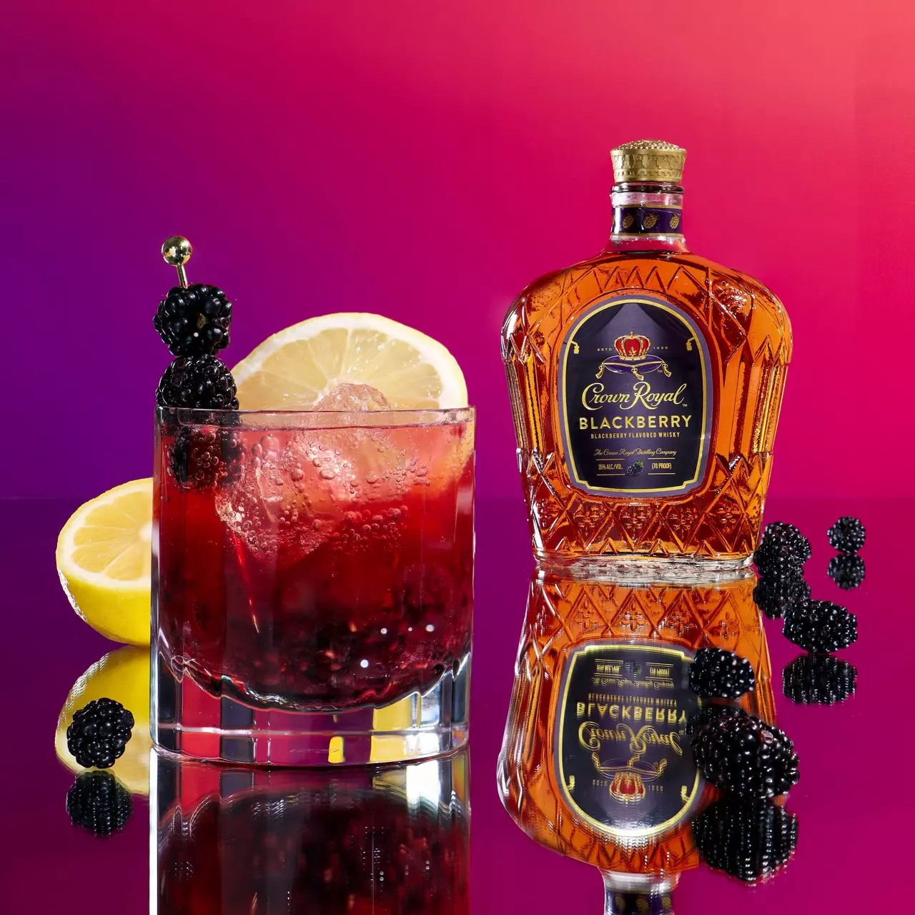A glass of blackberry cocktail with ice, lemon slice, and fresh blackberries sits next to a bottle of Crown Royal Blackberry whisky on a reflective surface with a purple-red gradient background.