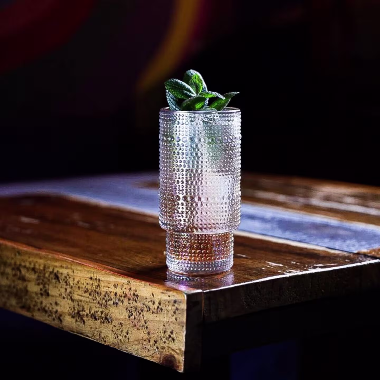 La imagen muestra un vaso de vidrio con un patrón texturizado de Love Cocktail Company, lleno de un cóctel transparentere, adornado con hojas de menta verde en la parte superior.