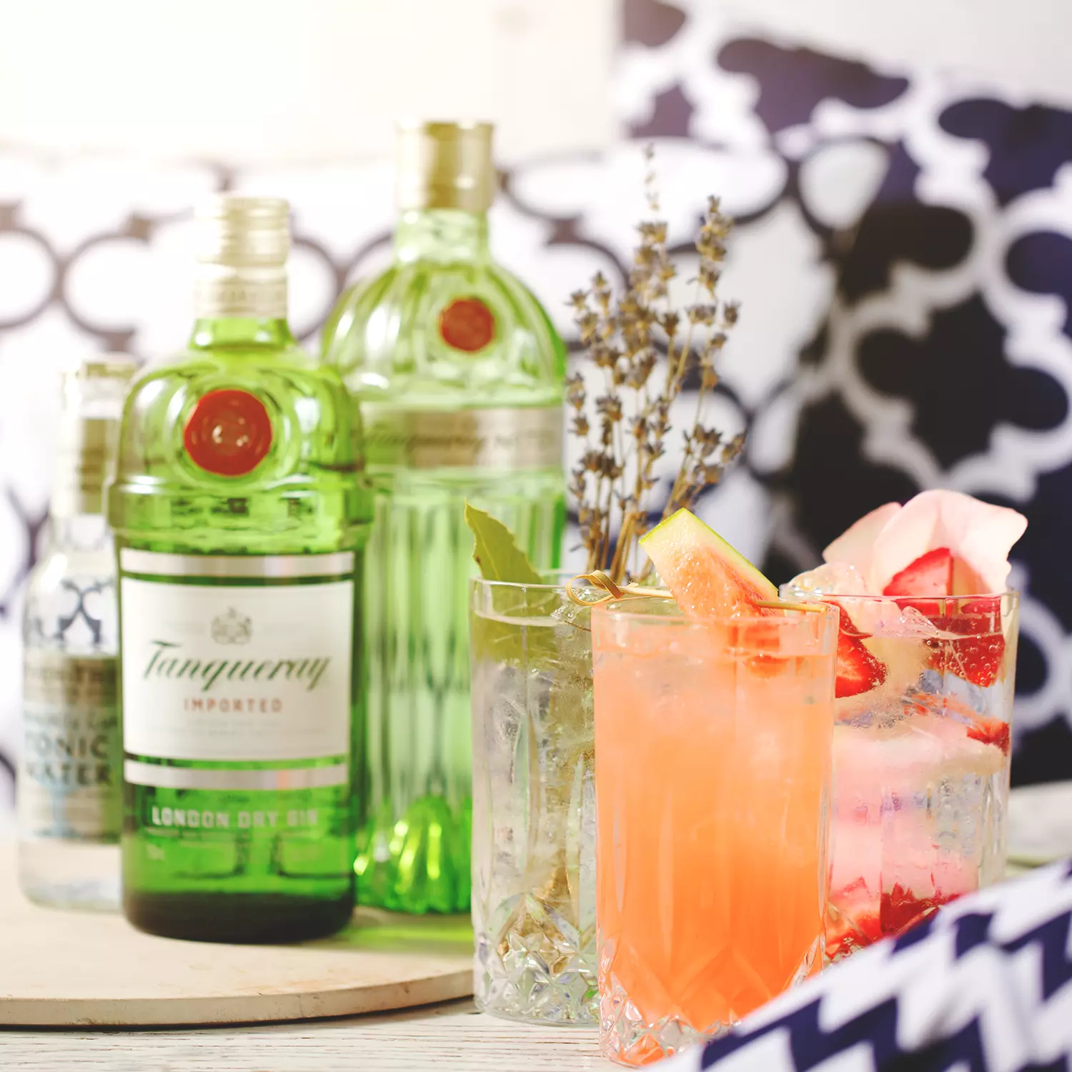 Two bottles of Tanqueray gin and a bottle of tonic water behind three colorful gin cocktails garnished with fruit and herbs, set on a table with patterned cushions in the background.