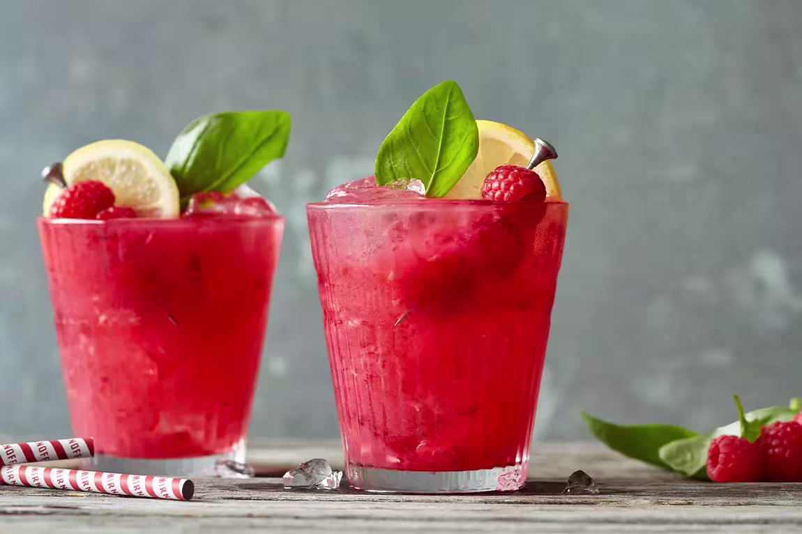 Two glasses of red raspberry drinks garnished with lemon slices, basil leaves, and whole raspberries, on a wooden surface with some ice and straws nearby.