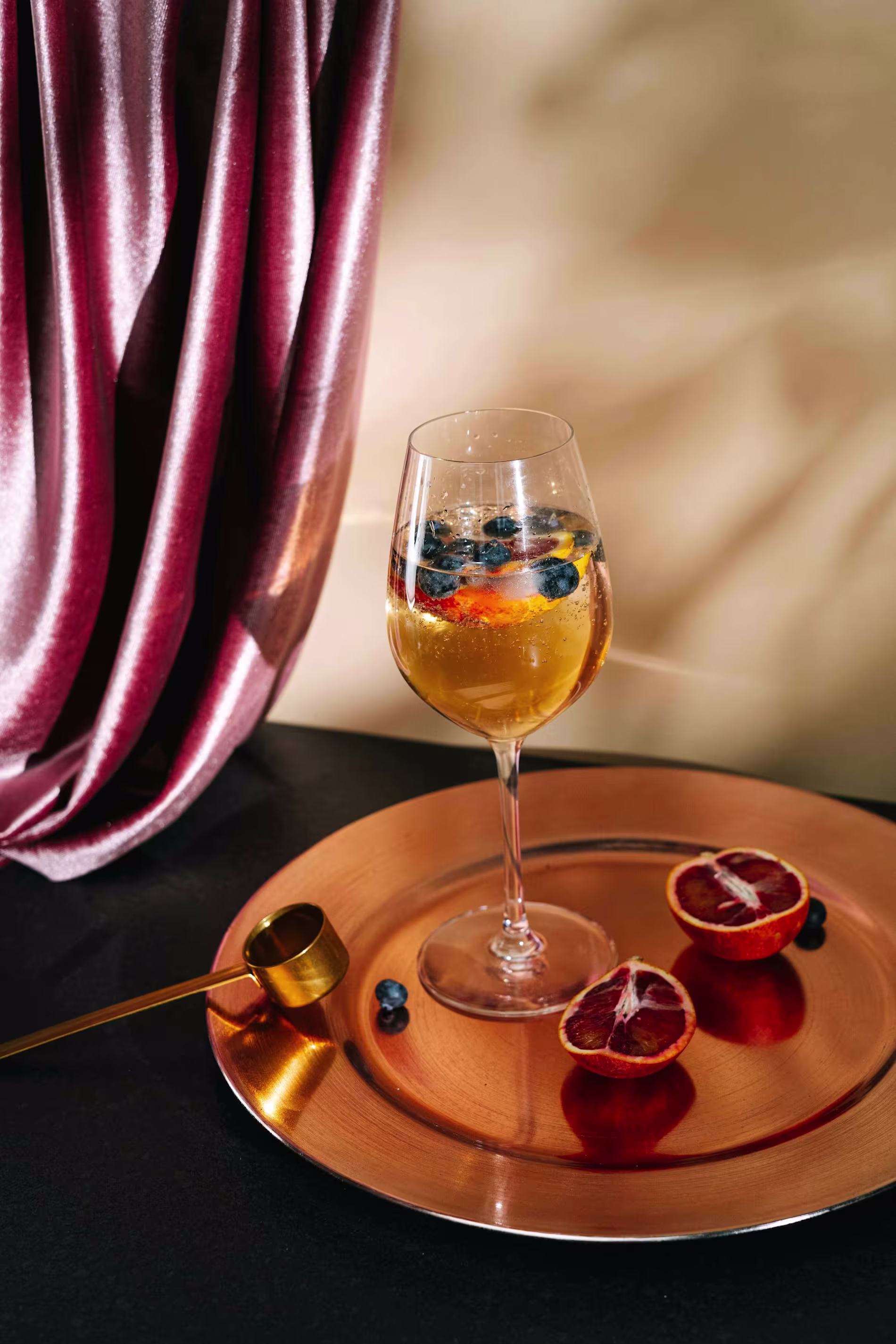A wine glass with a light-colored drink and berries, placed on a gold tray with a halved blood orange, a few blueberries, and a gold measuring jigger nearby.