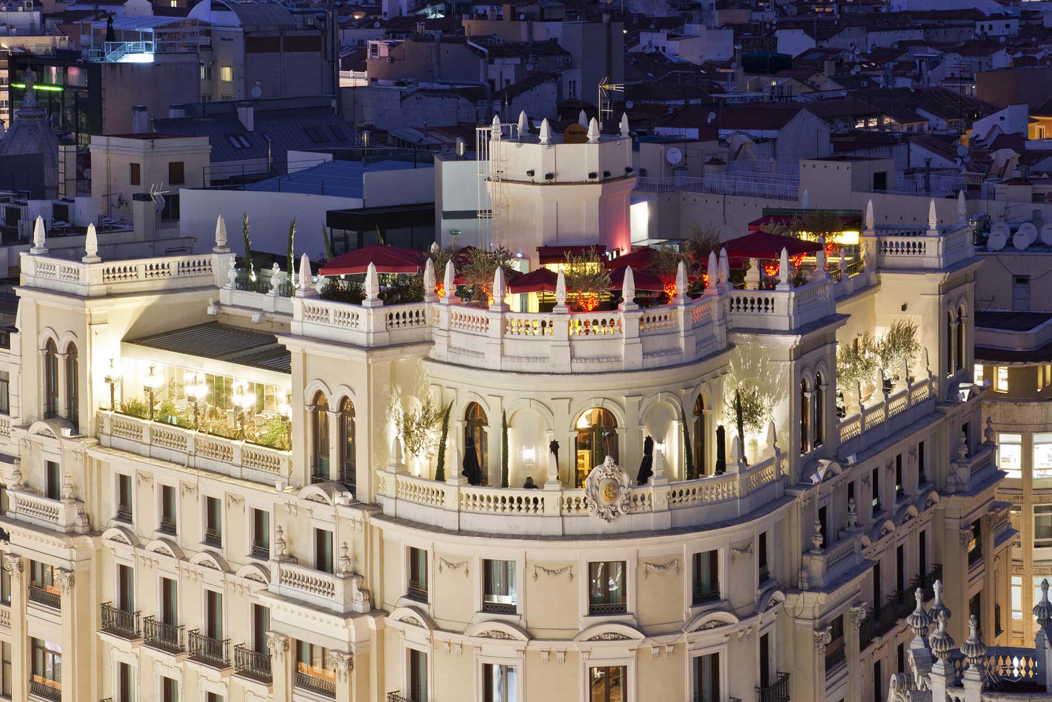Ático Restaurante y Terraza se muestra iluminado al anochecer, coronando un edificio clásico con detalles elegantes, sombrillas rojas y vegetación.