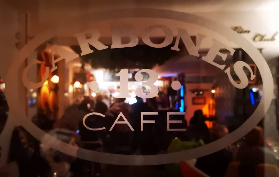 Carbones 13 refleja un ambiente acogedor a través de su cristal grabado, con luces cálidas y clientes disfrutando en el interior.