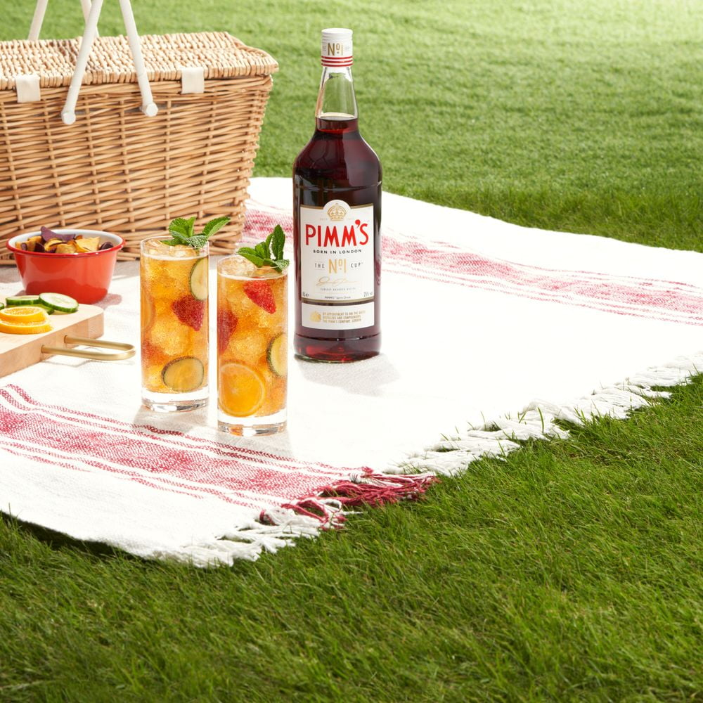 Pimm’s bottle with two glasses of Pimms cocktail garnished with mint, cucumber, and fruit on a picnic blanket next to a wicker basket
