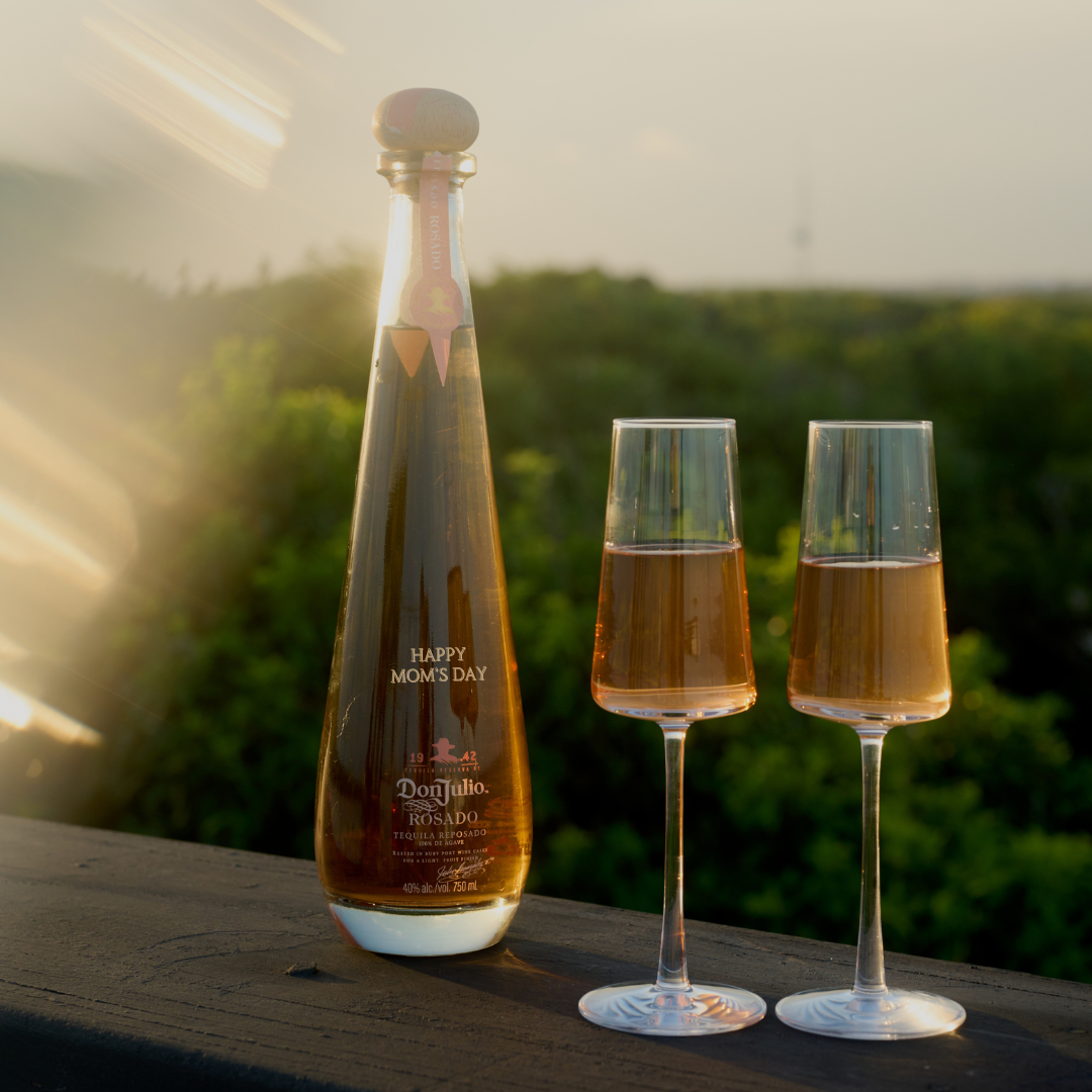A bottle of Don Julio Rosado tequila with Happy Moms Day on the label and two filled champagne flutes on a ledge outdoors, with greenery blurred in the background.