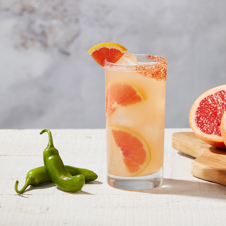 A Paloma cocktail with a grapefruit garnish next to two green jalapeno peppers.
