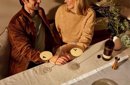 Two people sit at a table with cocktails and a bottle of liquor, smiling and holding hands. A jigger and vase with greenery are also on the table.